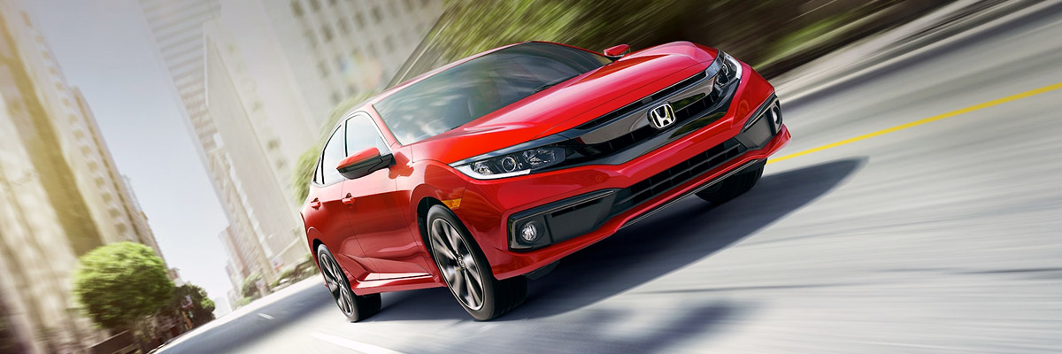 Front passenger-side view of the 2021 Civic Sport Sedan in Rallye Red driving on urban street with buildings in the background.