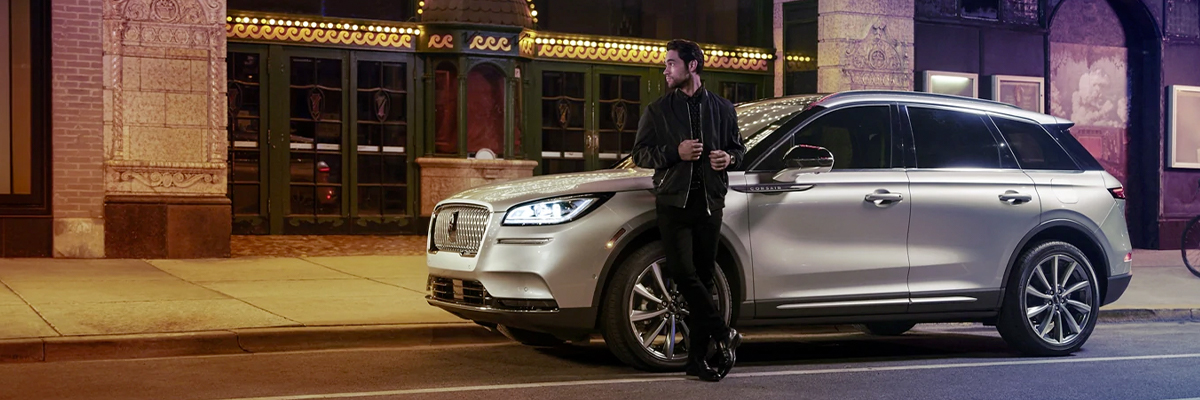 A 2021 Lincoln Corsair is parked outside a theatre as the driver relaxes against the frame and lights illuminate the floating roofline and body 