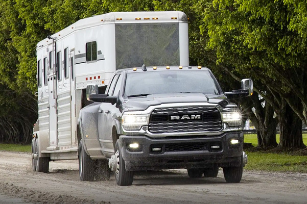 A 2021 Ram 3500 Limited towing a horse trailer, being driven on a dirt road next to a grove of large trees.