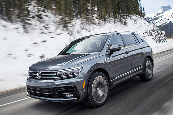 A Tiguan in Platinum Gray Metallic driving on a road as seen from the front with snowy mountains in the background.