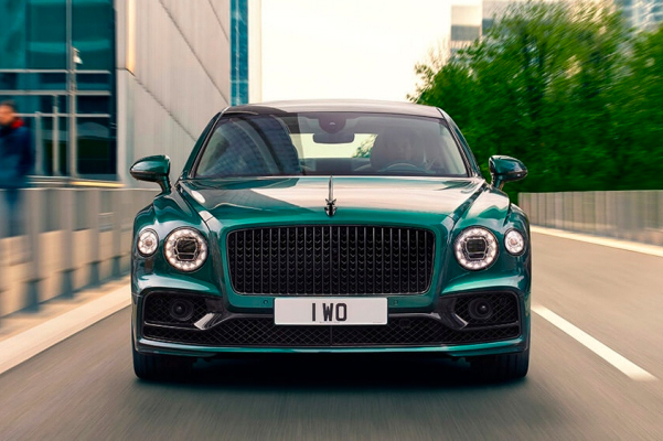 Exterior head on shot of a 2022 Bentley Flying Spur Hybrid driving with trees and buildings in the background.