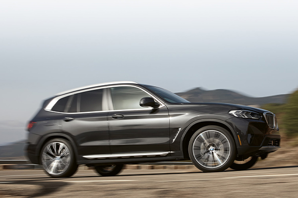 Exterior side shot of a 2022 BMW X3 driving up a road with mountains in the background.