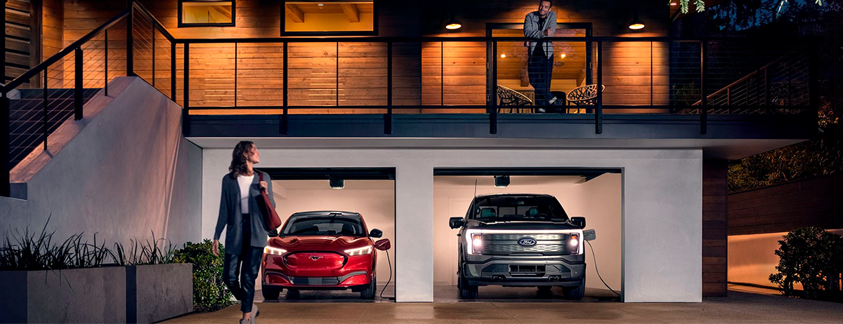 A 2022 Ford Mustang Mach-E® and a Ford F-150® LightningTM are being charged in a garage at night near two people