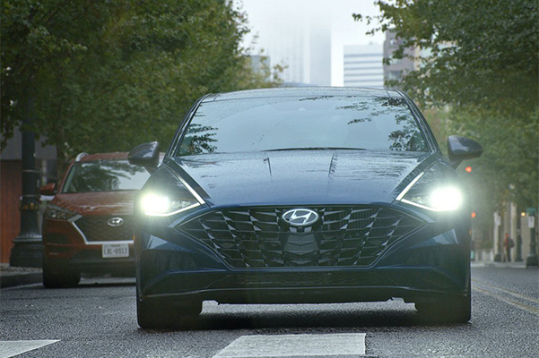 Front, exterior shot of a 2022 Hyundai Sonata driving down a city street on a hazy day with headlights on.