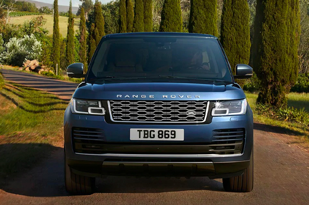Exterior head on shot of a 2022 Range Rover driving on a road with trees and a field in the background.