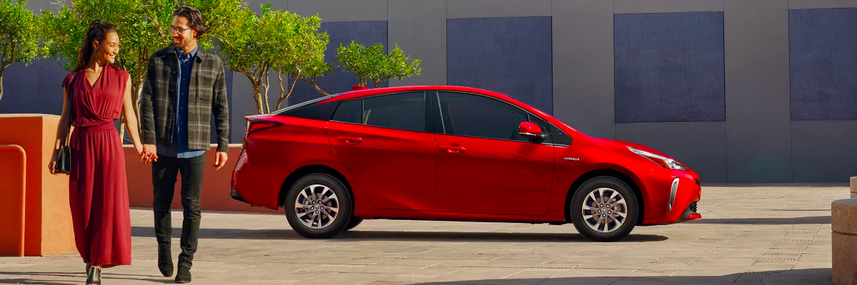 A 2022 Toyota Prius parked with trees and a building in the background and two people walking in front of the car.