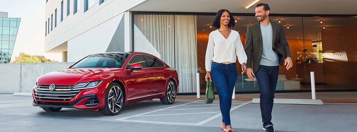 A smiling man and woman walk away from an Arteon shown in Kings Red Metallic, parked outside a modern building.