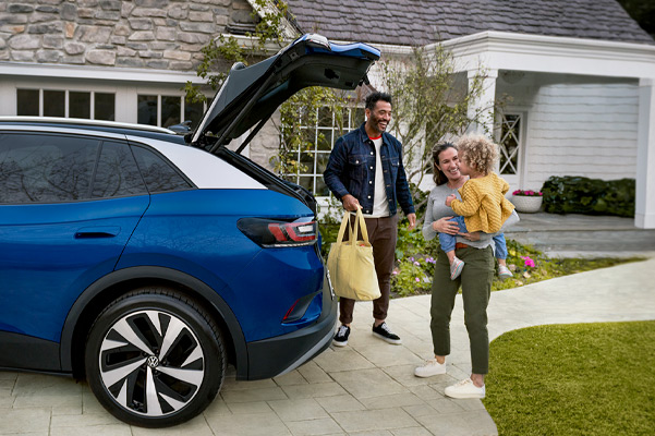 A young family loads a bag into the cargo area of an ID.4, shown in Dusk Blue Metallic parked in a residential driveway.
