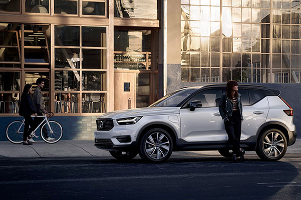 A woman stands leaning against a white XC40 Recharge plug in hybrid, behind her is a restaurant.
