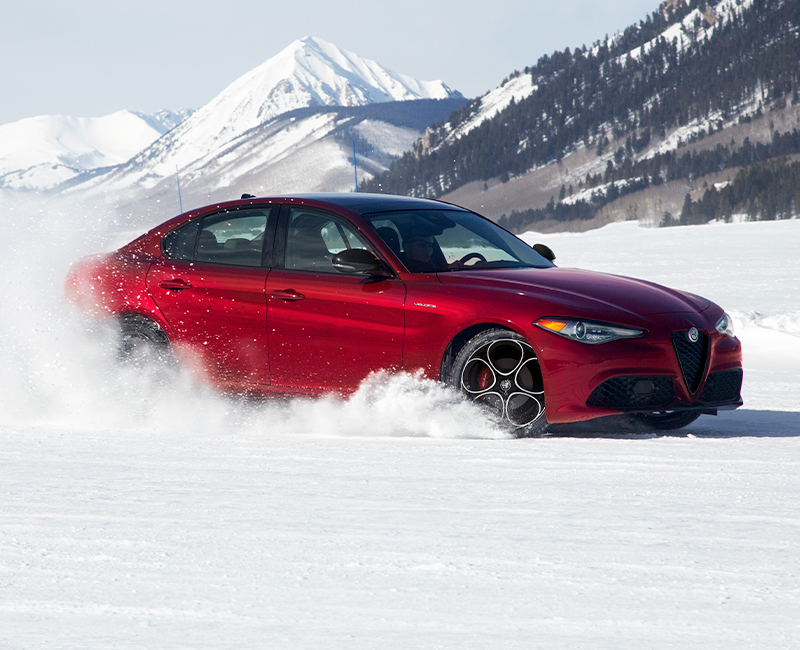 A red 2023 Alfa Romeo Giulia Veloce kicking up snow as it is being driven through the mountains.