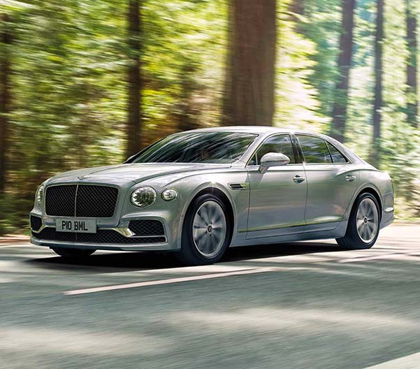Exterior shot of a 2023 Bentley Flying Spur Hybrid driving on a street with trees in the background.