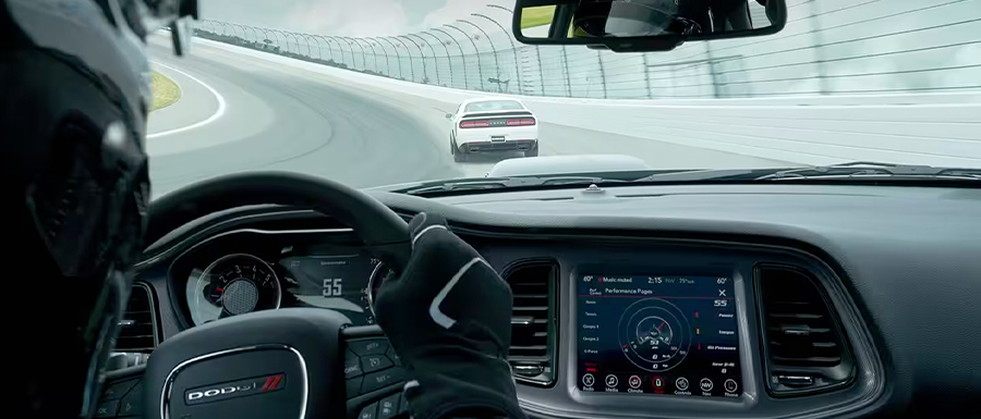 Display The interior of a 2023 Dodge Challenger as the driver takes a curve on a track, with the Digital Cluster displaying 55 mph and the Uconnect touchscreen displaying a Dodge Performance Page.