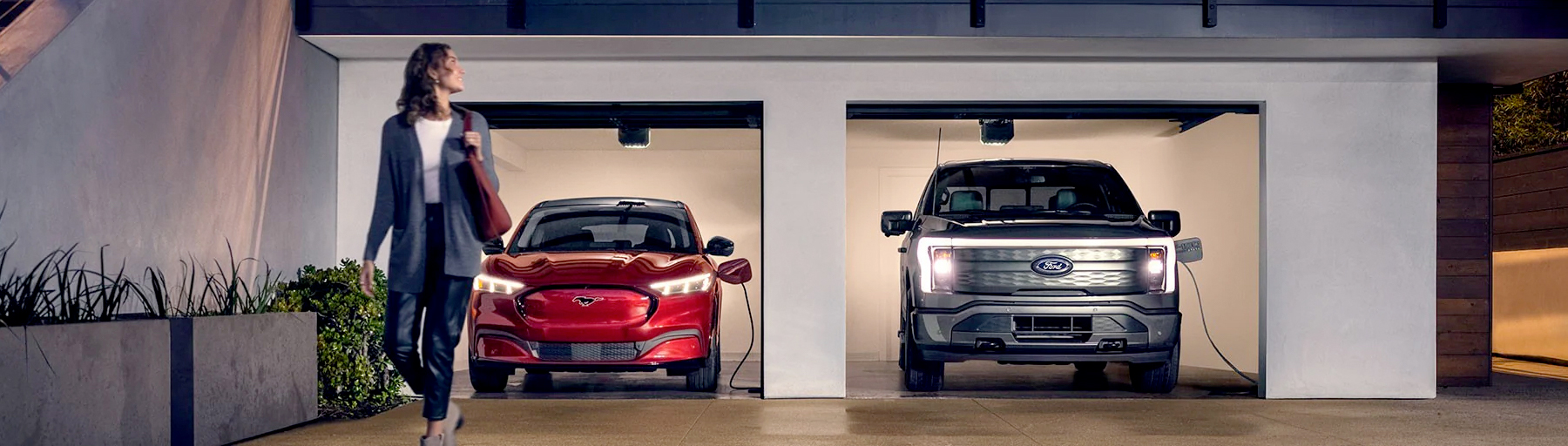 A 2022 Ford Mustang Mach-E® and a Ford F-150 Lightning are being charged in a garage at night near two people