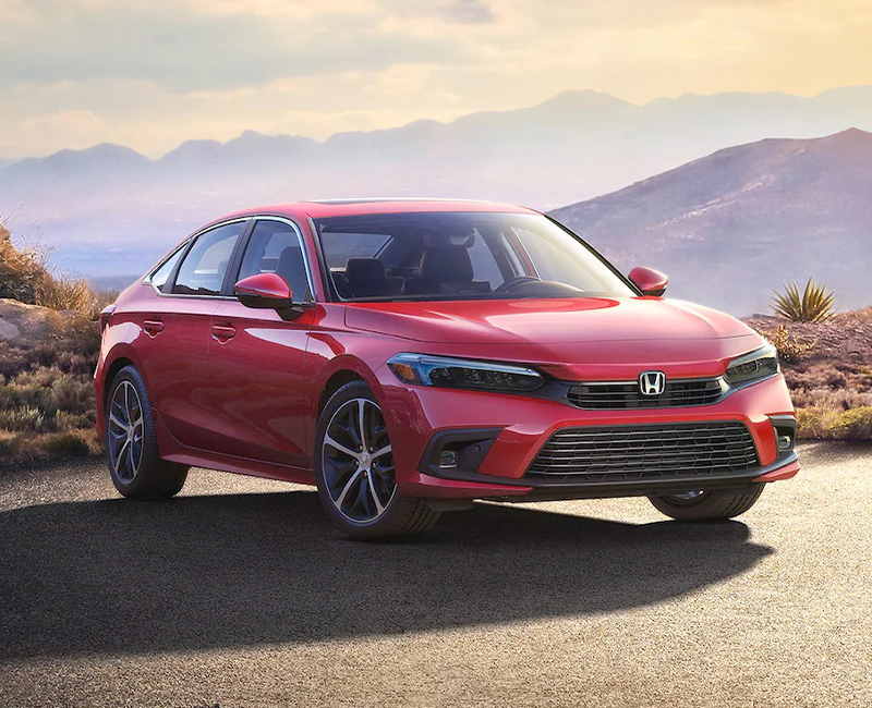 Front passenger-side view of the 2023 Honda Civic Touring Sedan in Rallye Red, parked in a misty parking lot at dusk with mountains in the background.