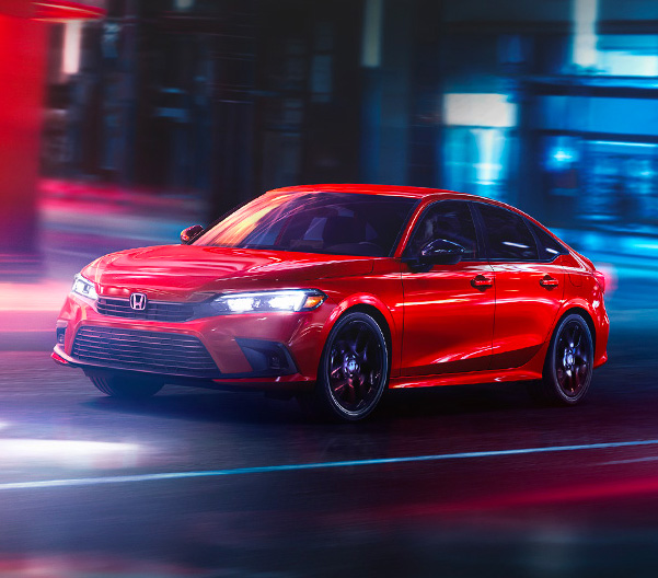 Front driver-side view of the 2022 Honda Civic Sport Sedan in Rallye Red, rounding a corner on a city street at night.