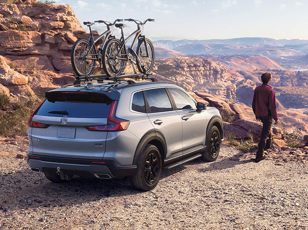 Rear passenger-side view of the 2023 Honda CR-V Sport Touring Hybrid in Lunar Silver Metallic with Honda Genuine Accessories, parked at a desert overlook with bikes on the roof attachment.