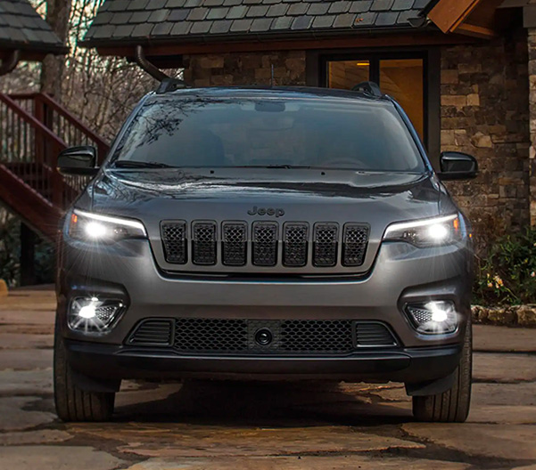 A head-on view of a 2023 Jeep Cherokee Altitude Lux with its headlamps and fog lamps on, parked in the driveway of a brick residential building.