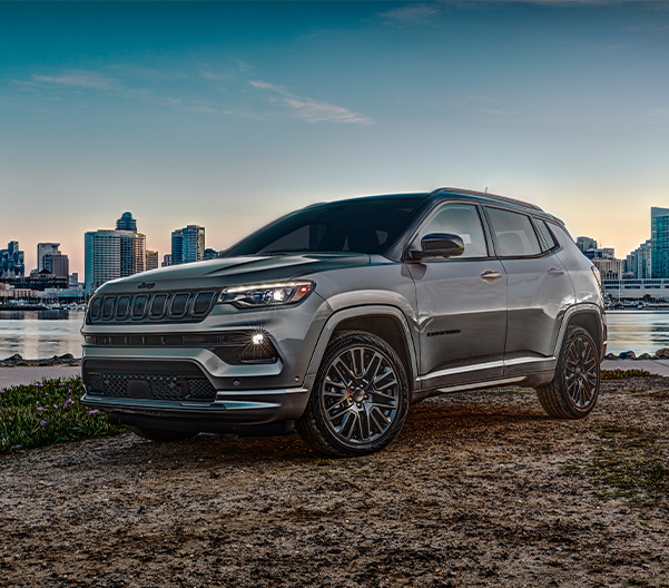 2022 Compass High Altitude in Billet Silver parked on sand near river with cityscape view at sunset.