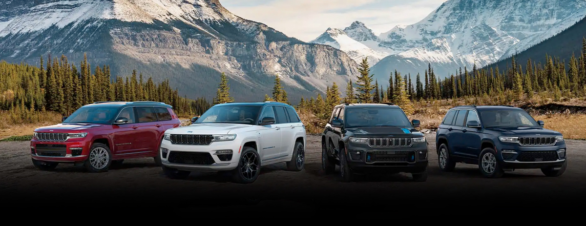 A lineup of four Jeep Grand Cherokee models parked on a clearing with snow-capped mountains in the background.