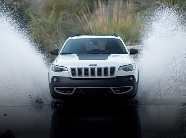 A head-on angle of a white 2023 Jeep Cherokee Trailhawk fording a stream off-road, with water splashing well above the top of the vehicle.