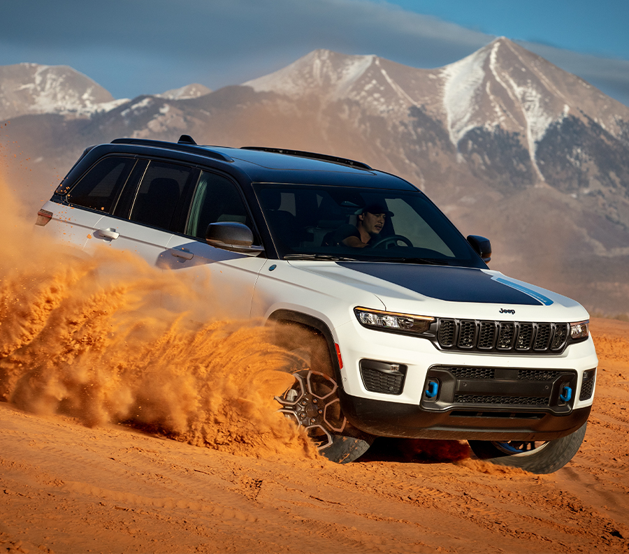 2023 Grand Cherokee Trailhawk 4xe in Bright White and Black kicking up a large cloud of sand while driving in the desert, with snow-capped mountains in the distance and grey clouds in the sky above.