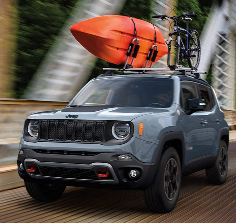 A gray 2023 Jeep Renegade Trailhawk with a kayak and bicycle strapped to its roof rack, being driven across a bridge with the background blurred to indicate the speed of the vehicle.