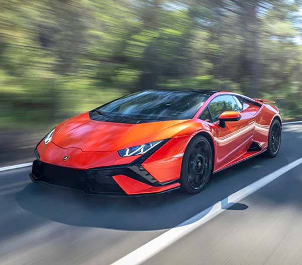 Exterior shot of an orange 2023 Lamborghini Huracán Tecnica driving down a road during the daytime with trees in the background.