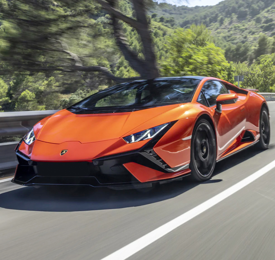 Exterior shot of a 2023 Lamborghini Huracán driving on a road with trees and mountains in the background.