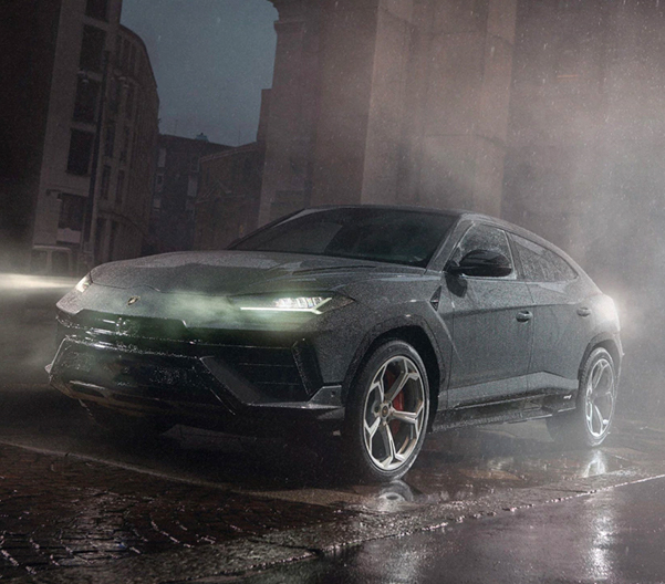 An exterior shot of a New Lamborghini Urus S driving with the headlights on during a rainy night in a city.