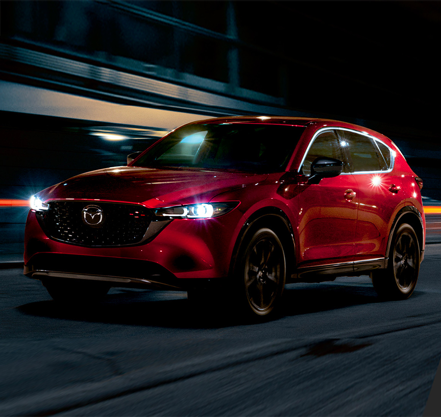Red 2023 Mazda CX-5 driving down street at night with lights on.