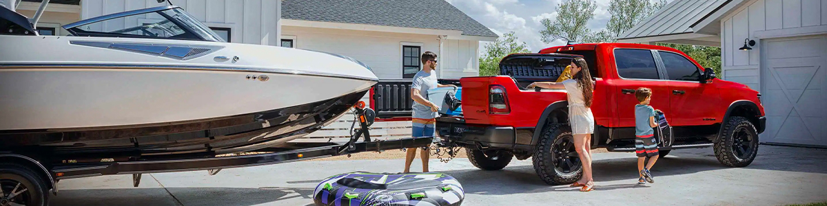 A 2023 Ram 1500 with a motorboat in tow and a man and woman loading a wakeboard and floaties into the truck bed and the RamBox.