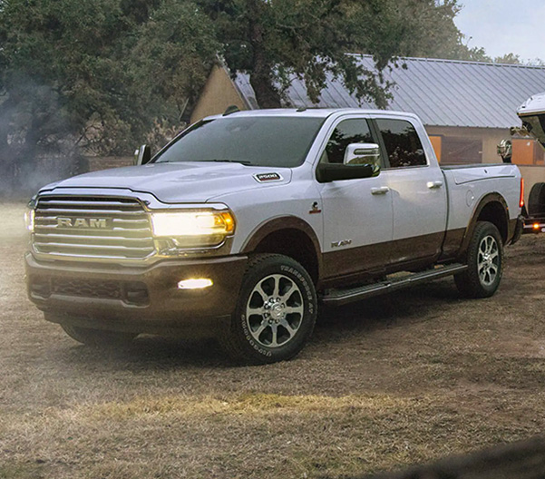 Display A white 2023 Ram 2500 Limited Longhorn Crew Cab with its headlamps on, towing a large speedboat on a dirt road.