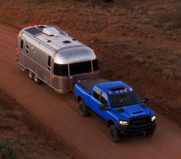 Display A view from above of a blue 2023 Ram 2500 Power Wagon Crew Cab with its headlamps on, towing a travel trailer as it is driven down a dirt road.