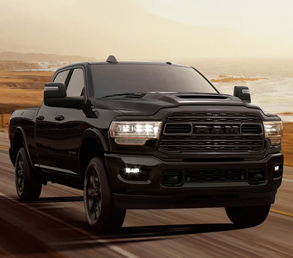 A 2023 RAM 3500 Limited Crew Cab with its headlamps on, being driven on a waterfront highway at dusk.