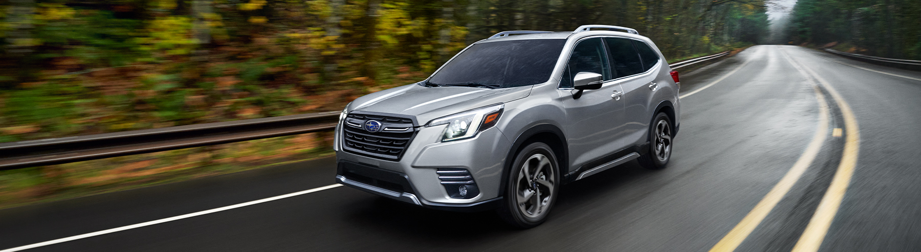 A silver Subaru Forester small SUV drives on a road past a wooded area