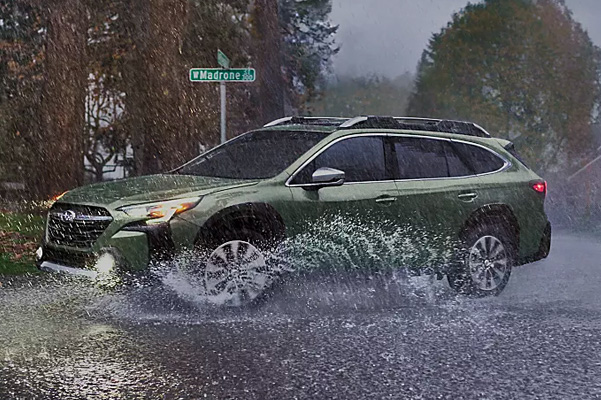 Exterior shot of a 2023 Subaru Outback driving through a puddle on a street corner.