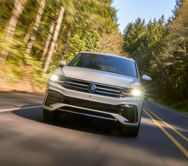 A Tiguan shown in Opal White, driving on a two-lane road with trees in the background, as seen from front.