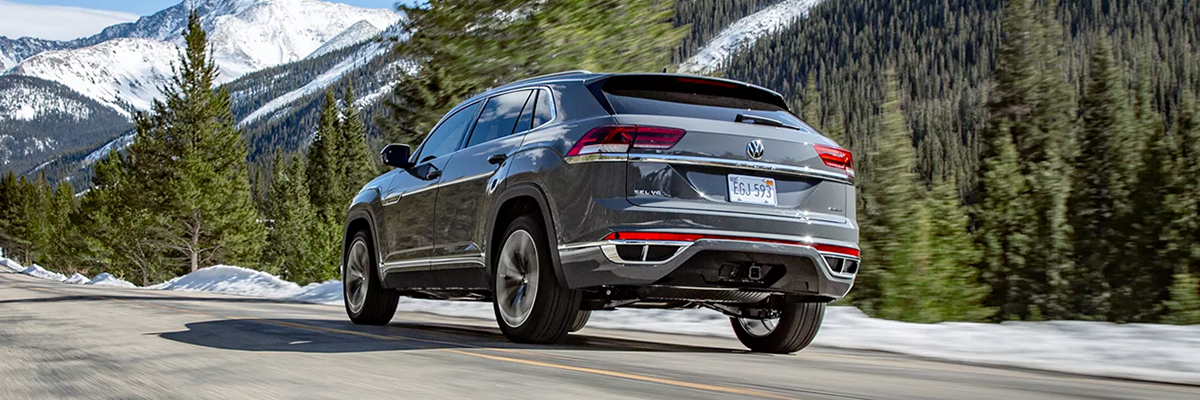 A low-angled shot looking at an Atlas Cross Sport shown in Pure Gray driving away, up a hilly road toward snow-covered, mountains.