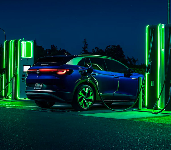 An ID.4 in Dusk Metallic Blue as it is parked at night next to a neon lit charging station.