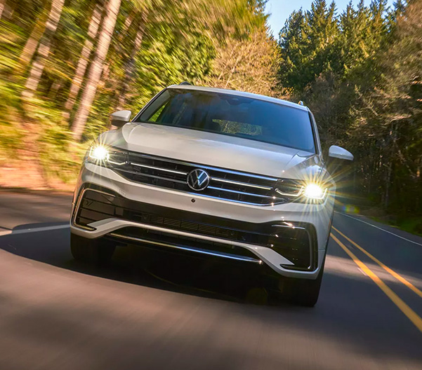 A Tiguan shown in Opal White, driving on a two-lane road with trees in the background, as seen from front.