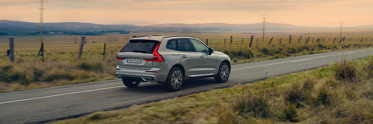 The 2023 Volvo XC60 driving away on a street surrounded by grassy fields and mountains in the background