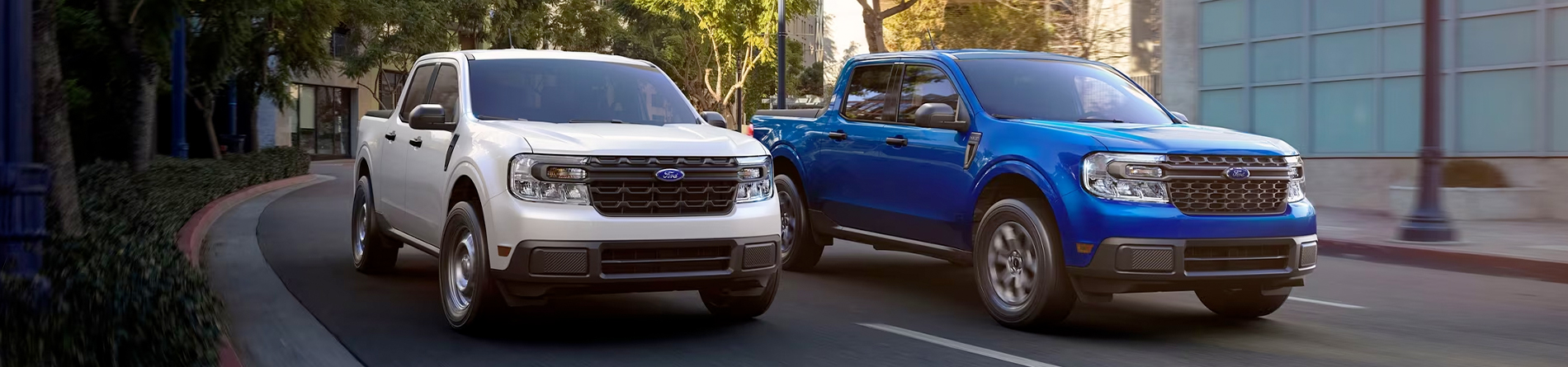 Two 2024 Ford Maverick® trucks in Oxford White and Atlas Blue Metallic are being driven on a tree-lined city road