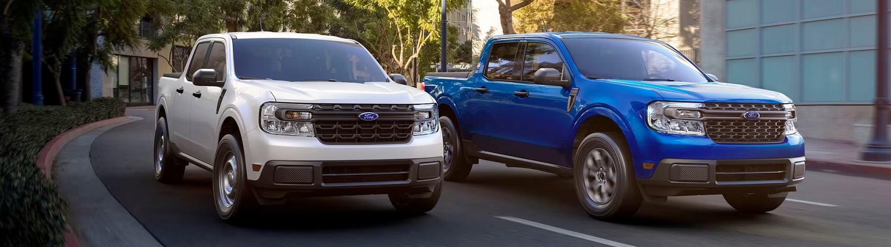 Two 2024 Ford Maverick® trucks in Oxford White and Atlas Blue Metallic are being driven on a tree-lined city road