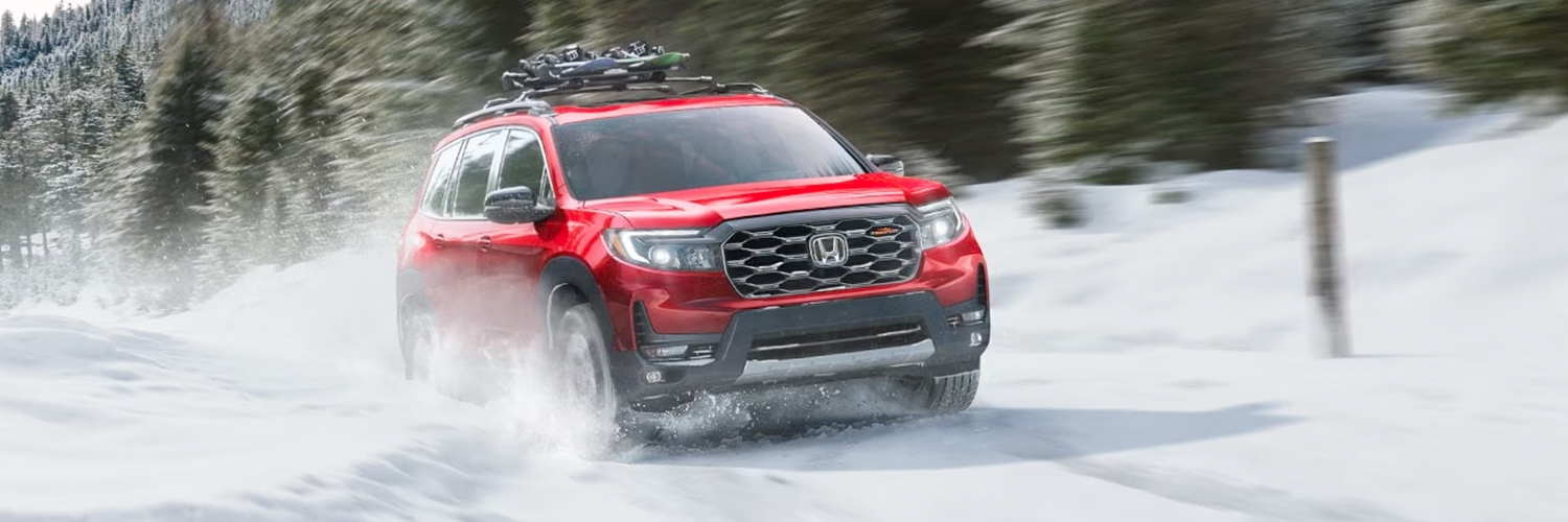 Front passenger-side view of the 2024 Honda Passport TrailSport with Honda Genuine Accessories, driving over a snow-covered road in a wilderness environment.