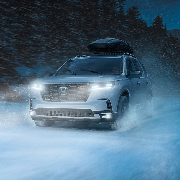 Honda Pilot shown with accessory crossbars and roof box driving at night in the snow.