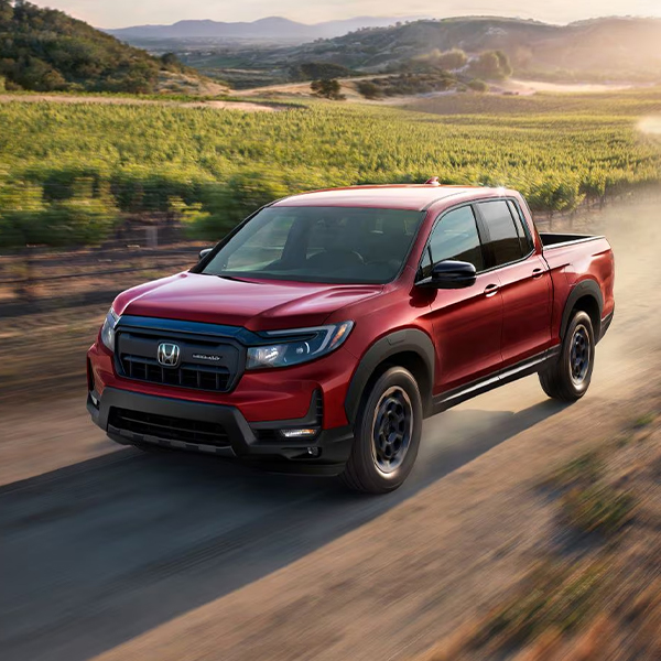 2024 Honda Ridgeline Sport in Radieant Red Metallic II driving on a dirt road through a vineyard