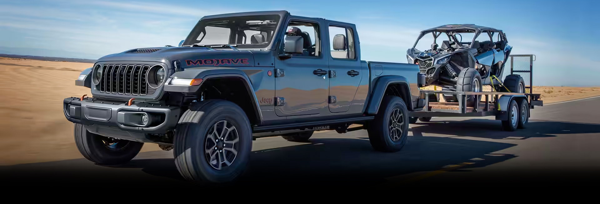 A gray 2024 Jeep Gladiator Mojave being driven down a highway as it tows an ATV on a utility trailer.
