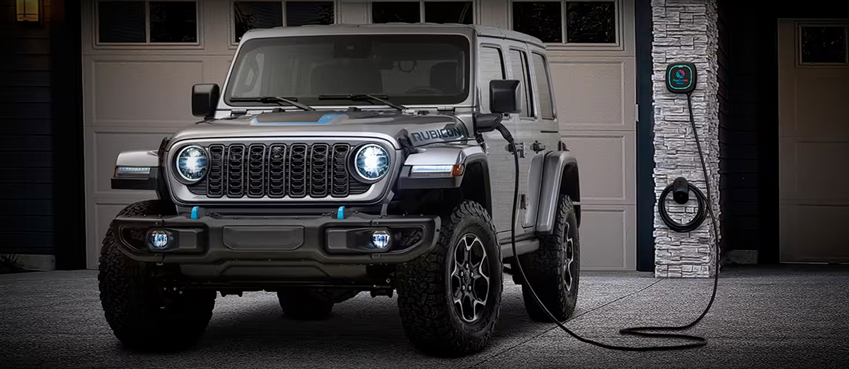 A 2024 Jeep Wrangler Rubicon 4xe parked in front of a garage with a built-in charging station and its charging cord plugged into the vehicle's charging port.