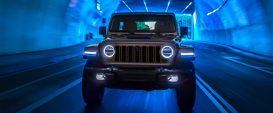 A front angle of a gray 2024 Jeep Wrangler Rubicon 4xe with its headlamps on, being driven through a lighted tunnel, with the entire image tinted blue.
