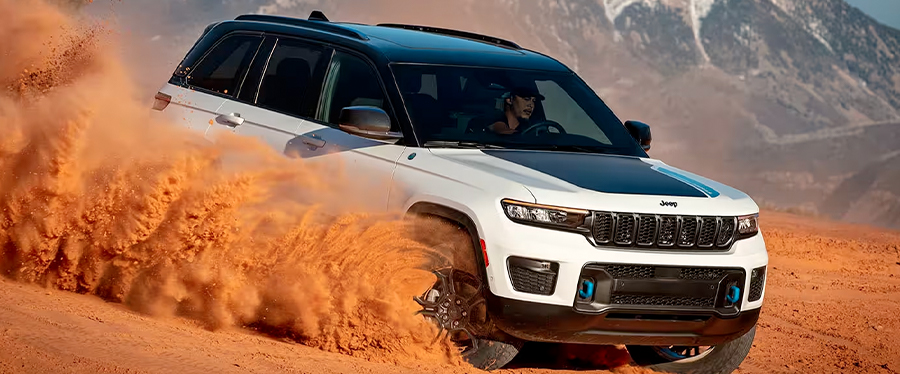 A white 2024 Jeep Grand Cherokee Trailhawk 4xe kicking up a cloud of orange sand and dust as it's being driven down a dune, with mountains in the background.